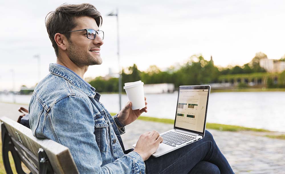 Junge sitzt auf einer Bank, trinkt einen Kaffee und schaut ruhig auf seinen Computer